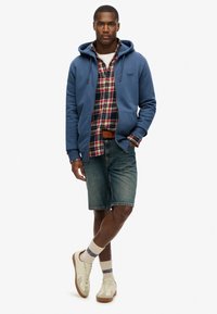 Sudadera con capucha azul sobre una camisa de cuadros, pantalones cortos de denim, zapatillas blancas y beige, y calcetas con estampado. Atuendo casual en capas.