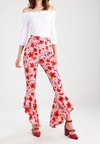 Witte off-shoulder top met smockdetails, gecombineerd met roze flare-broek met rode bloemenpatronen en rode hoge hakken.