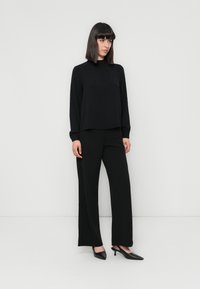 Black long-sleeve top with subtle texture, paired with wide-leg black trousers. The model wears black pointed-toe heels. Simple design and color.
