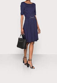 Mörkblå klänning med korta ärmar och åtsittande midja, accentuerad med ett silverbälte. Kombinerad med svarta högklackade sandaler och en handväska.