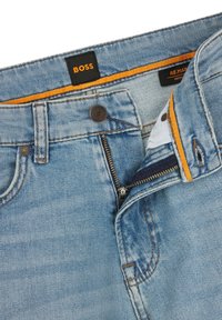 Jeans de mezclilla azul claro con etiqueta BOSS y costuras amarillas, mostrando una cremallera parcialmente abierta en la parte delantera y un dobladillo limpio.