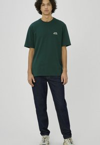 Jeune personne aux cheveux bouclés portant un t-shirt vert foncé avec un texte, un jean foncé et des baskets blanches, debout devant un fond uni.