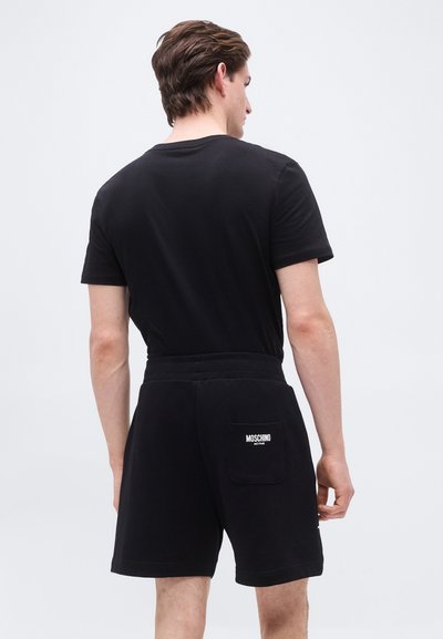 Άνδρας φορώντας μαύρο κοντομάνικο πουκάμισο και μαύρο σορτς με το λογότυπο Moschino Active στην πίσω τσέπη, στέκεται μπροστά σε απλό φωτεινό φόντο.