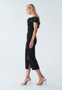 Black off-shoulder dress with a ruffled neckline and a front slit, fitted shape, and knee-length hem. Paired with beige high-heeled sandals.