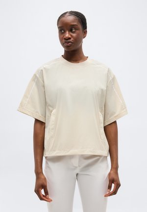 Γυναίκα με πλεξούδα, φορώντας ανοιχτό, κοντομάνικο μπλουζάκι μπεζ και λευκό παντελόνι, που στέκεται μπροστά σε απλό λευκό φόντο.