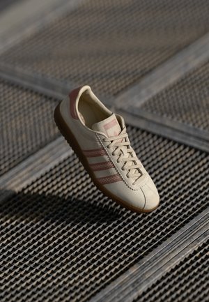 Beige atletik sko med røde aksenter, tre-stripe design, teksturert overflate og gummisåle, vist på et mønstret metallunderlag.
