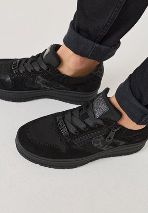Baskets basses noires avec fermeture éclair latérale, détails imprimé léopard et lacets noirs portées avec un pantalon noir retroussé sur un sol blanc.