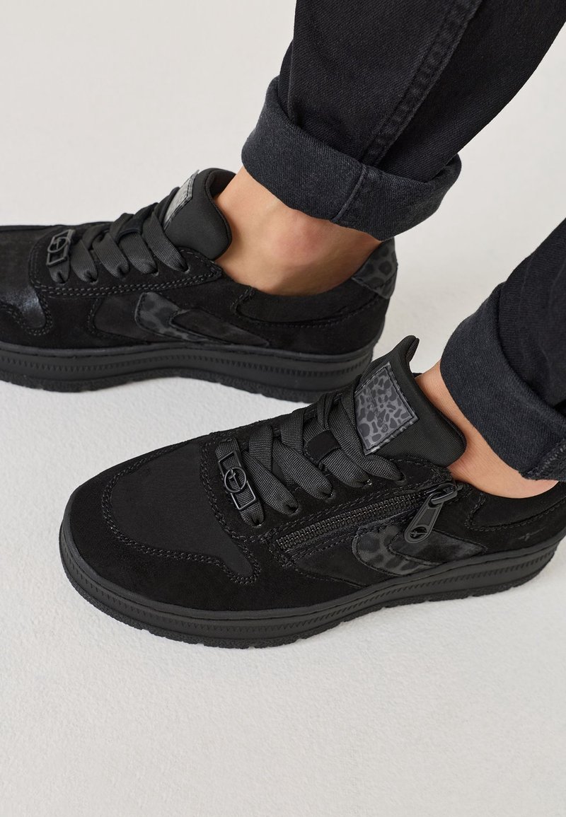 Baskets basses noires avec fermeture éclair latérale, détails imprimé léopard et lacets noirs portées avec un pantalon noir retroussé sur un sol blanc.