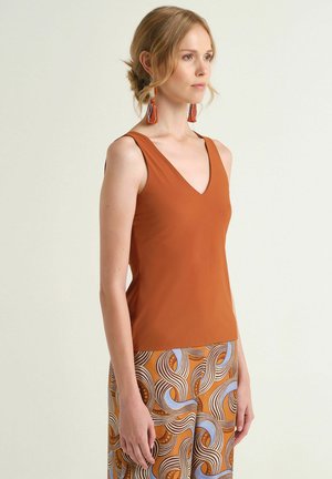 Giovane donna che indossa un top senza maniche color ruggine, pantaloni con motivi arancioni, marroni e blu, e orecchini con nappine coordinati, in piedi di lato.