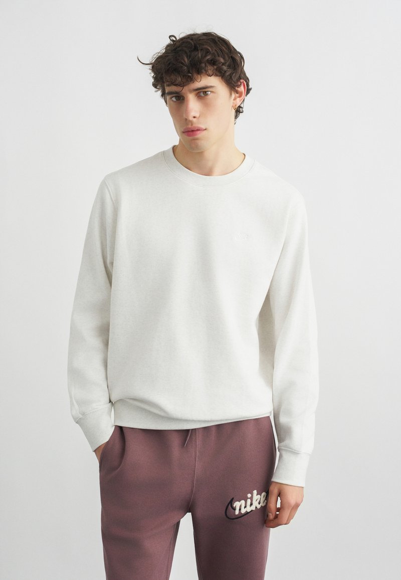 Giovane uomo con capelli ricci che indossa una felpa bianca e pantaloni della tuta Nike color malva, in piedi davanti a uno sfondo chiaro uniforme.