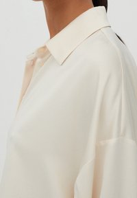 Camicia beige con bottoni, colletto classico, in tessuto leggero e vestibilità rilassata. Texture liscia con una leggera lucentezza e morbida caduta.