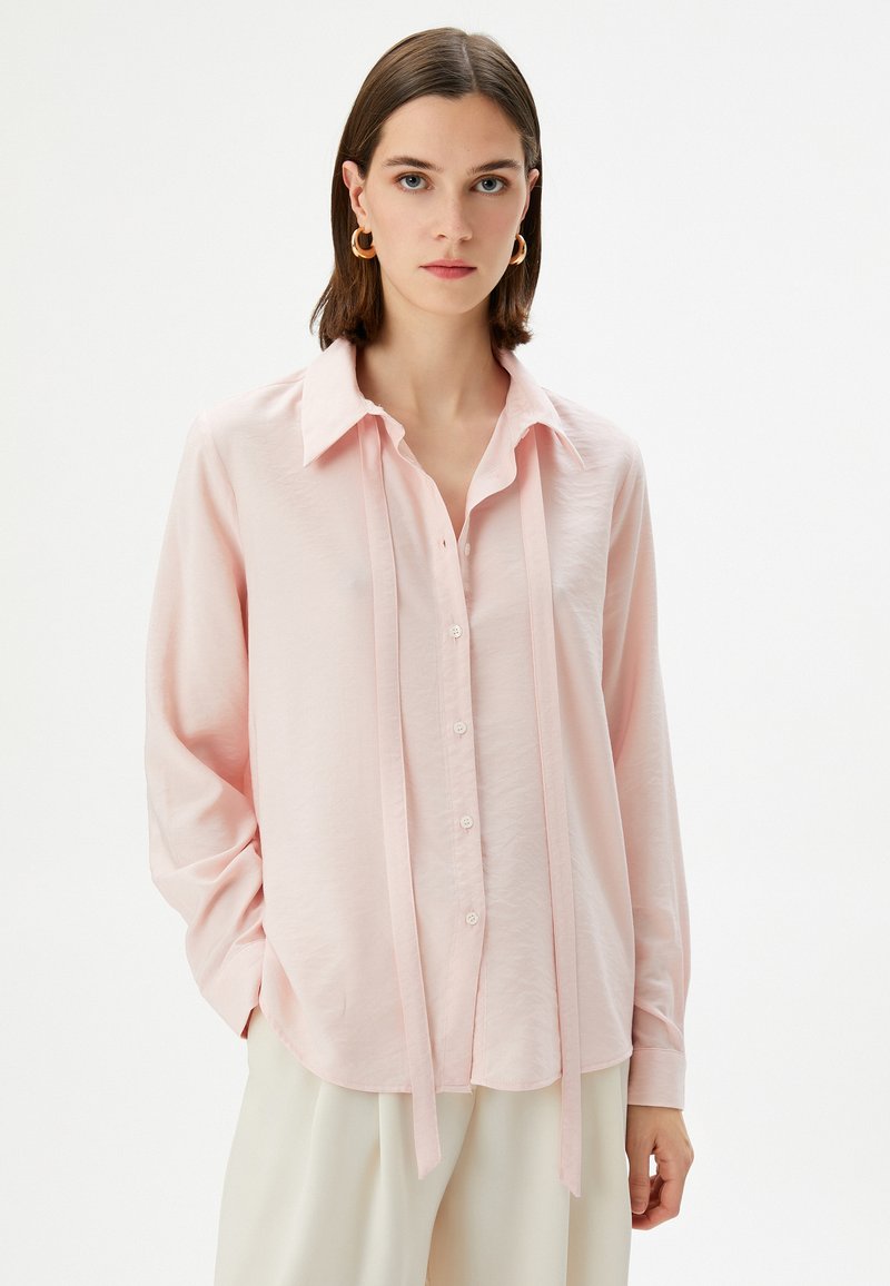 Lichtroze blouse met lange mouwen, een kraag, knoopsluiting en strikjes bij de hals. Gemaakt van gladde, lichte stof.