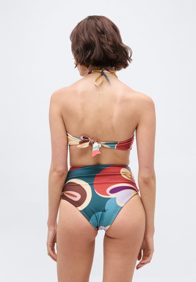 Γυναίκα με κοντά καστανά μαλλιά που φοράει πολύχρωμο ψηλόμεσο μπικίνι με αφηρημένα μοτίβα, ορατή από πίσω πάνω σε ένα απλό φόντο.