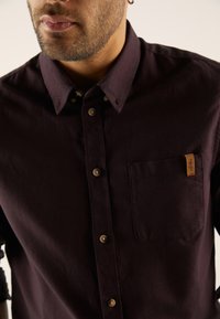 Dark burgundy button-up shirt made of smooth fabric, featuring a pocket with a leather label and subtle buttons on the collar.