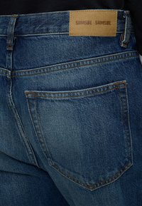 Gros plan sur la poche arrière d’un jean en denim bleu avec une étiquette de marque en cuir marron portant l’inscription "Samsøe Samsøe" sur la ceinture.