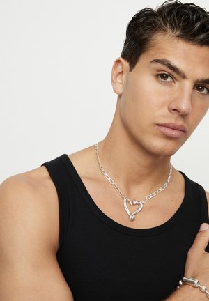 Collar de cadena de plata con un colgante en forma de corazón, llevado sobre una camiseta sin mangas negra, que muestra una textura suave y un diseño de corazón abierto.