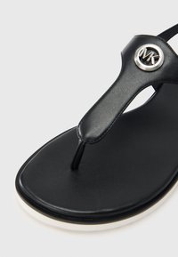 Μαύρα σανδάλια με λουρί από λείο δέρμα και κυκλικό ασημί λογότυπο. Η σόλα είναι επίπεδη με αντίθετη λευκή άκρη.