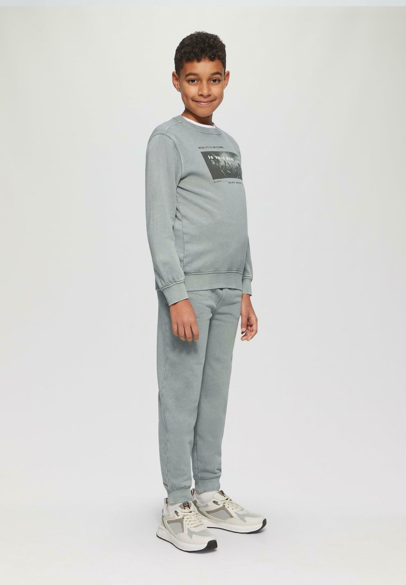 Lichtgrijze katoenen sweatshirt met zwart grafisch ontwerp, gecombineerd met bijpassende lichtgrijze broek. Schoenen hebben een witte basis met grijze accenten.