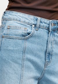 Ανοιχτό μπλε τζιν παντελόνι με ασημένια νύχια και ραφές, φορεμένο με καφέ τοπ που φαίνεται στη μέση.