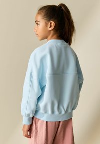 Sweat-shirt bleu clair avec un col rond, une coupe ample et des poignets froncés. Associé à un pantalon texturé rose pâle. Finition en tissu lisse.