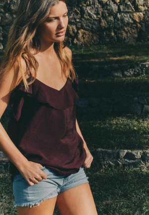 Noor naine tumelilla varrukateta topis ja hele denimsärgiga seisab õues kiviastmete ja rohu lähedal.