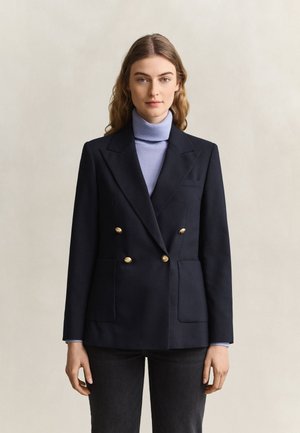 Femme portant un blazer bleu marine croisé avec des boutons dorés sur un pull à col roulé bleu clair et un pantalon foncé, debout devant un fond uni.