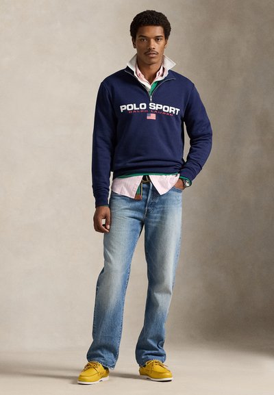 Μπλε σκούρο φούτερ με λογότυπο "POLO SPORT", φορεμένο πάνω από ροζ πουκάμισο με κουμπιά. Το σύνολο ολοκληρώνουν ανοιχτό γαλάζιο τζιν και κίτρινα παπούτσια.