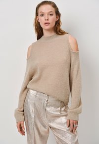 Beige, cold-shoulder sweater met een zachte textuur en hoge halslijn, gecombineerd met glanzende zilveren broek. Relaxte pasvorm met lange mouwen.