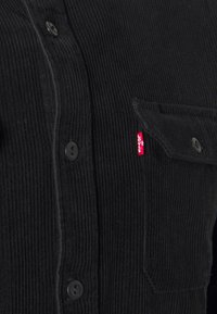 Chemise en velours côtelé noire avec un devant boutonné, deux poches poitrine et une étiquette Levi's rouge sur la poche gauche. Tissu texturé avec des côtes verticales.