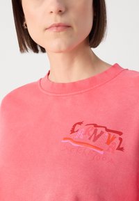Sudadera rosa con cuello redondo y un diseño gráfico en rojo y rosa. El material parece suave con una textura lisa.