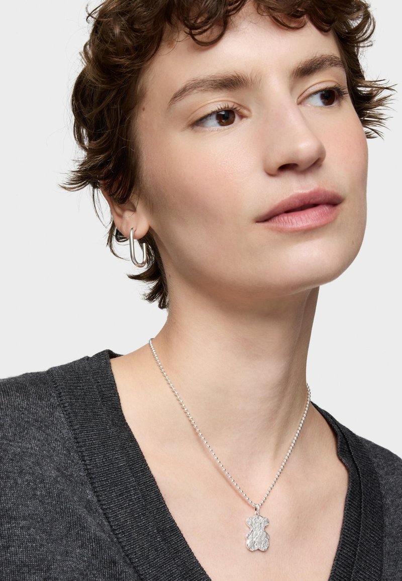 Collar con colgante de oso plateado sobre una cadena delicada, llevado sobre un suéter texturizado gris oscuro. El colgante presenta un acabado martillado.