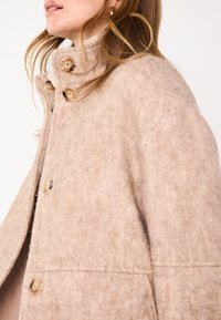 Cappotto beige in misto lana con colletto alto, dotato di grandi bottoni rotondi e una finitura morbida e testurizzata. Design strutturato con cuciture laterali.