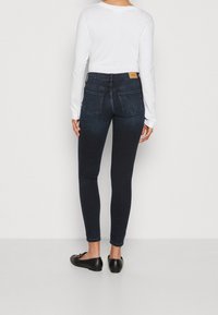 Vaqueros oscuros de denim, ajustados y de tiro alto, con diseño de cinco bolsillos y un ligero desgaste, combinados con una blusa blanca de manga larga.