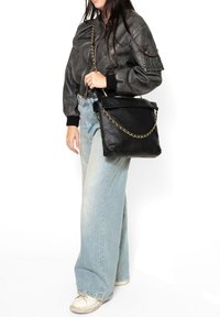 Borsa a tracolla in pelle nera trapuntata con chiusura a pattina e tracolla in catena dorata, abbinata a una giacca bomber grigia e jeans azzurri chiari.