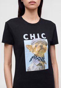Γυναίκα που φοράει μαύρο T-shirt με ένα σκύλο που φοράει καπέλο, τζιν μπουφάν και πουά μαντήλι, με τη λέξη "CHIC" πάνω από τη φωτογραφία του σκύλου.