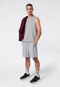 Top grigio chiaro senza maniche, pantaloncini grigi abbinati e sneakers nere. La modella tiene in mano una giacca color bordeaux e indossa calzini a righe con un logo.