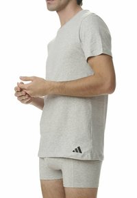 Mężczyzna w jasnoszarej koszulce Adidas i pasujących majtkach, stoi z plecami skrzyżowanymi na białym tle.