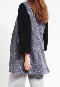 Vrouw met lang bruin haar, die een mouwloze grijze, gestructureerde vest draagt over een zwarte langmouwtop en lichtgrijze broek, staand in zijaanzicht.