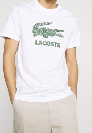 Camiseta blanca de algodón con un logo de cocodrilo verde y la palabra "LACOSTE" debajo, mangas cortas y diseño clásico de cuello redondo.