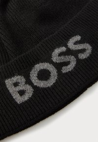 Zwarte gebreide beanie met het woord "BOSS" in contrasterend grijs garen, voorzien van een ribbelstructuur en een rond bovenontwerp.