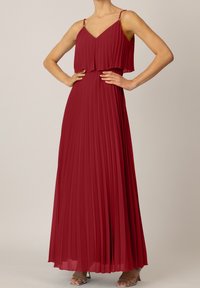 Femme portant une robe rouge longue plissée, sans manches, à fines bretelles, et des sandales à talons hauts argentées, les mains sur les hanches.