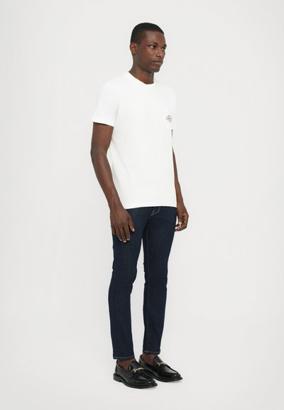 Camiseta blanca de manga corta con un bolsillo en el pecho que presenta un logo colorido, combinada con jeans ajustados azul oscuro y mocasines negros.