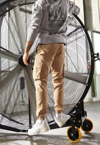 Pantaloni cargo marroni, felpa grigia con grafica e sneakers bianche. In piedi vicino a un grande ventilatore circolare con pale metalliche.
