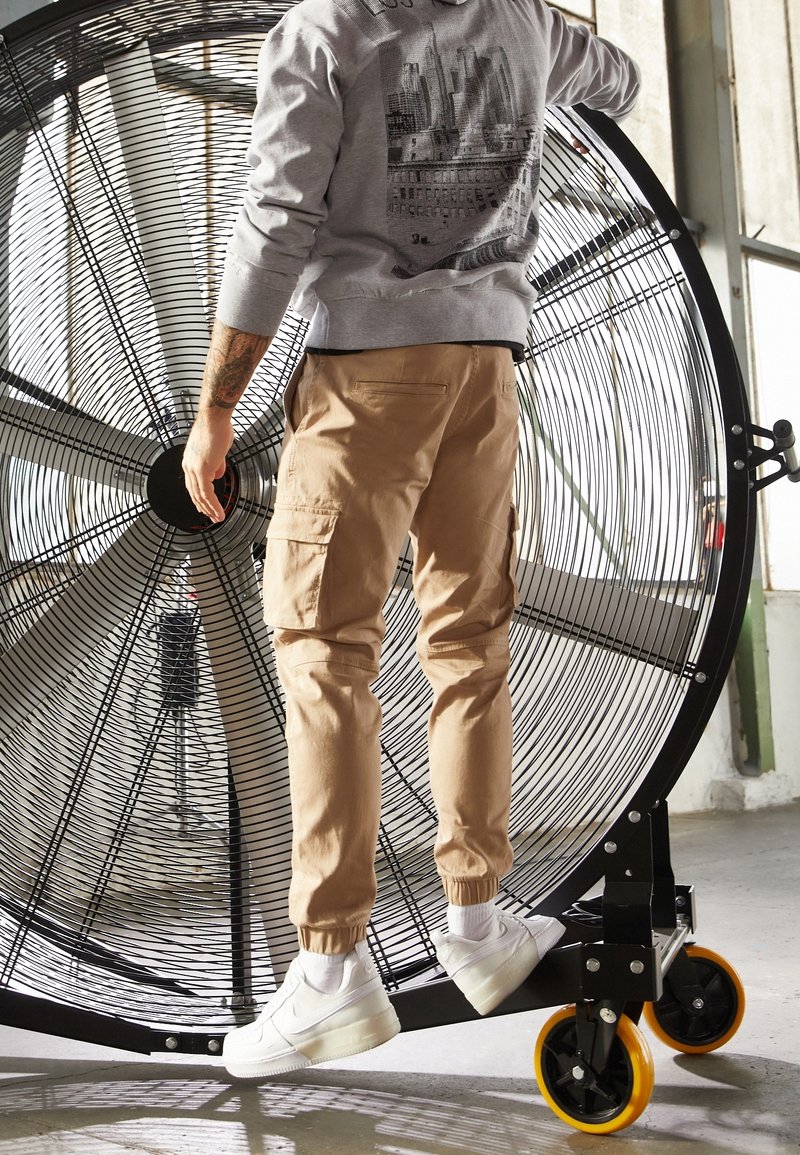 Pantaloni cargo marroni, felpa grigia con grafica e sneakers bianche. In piedi vicino a un grande ventilatore circolare con pale metalliche.
