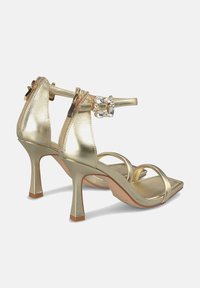 Goldene, metallische High-Heelsandalen mit einem Knöchelriemen, quadratischer Zehenpartie und kristallenen Verzierungen am Riemen. Glatte Textur und glänzende Oberfläche.