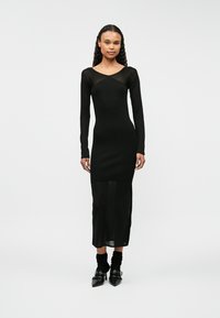 Czarna dopasowana sukienka z długimi rękawami, dekoltem w kształcie litery V i przezroczystym dolnym wykończeniem. Gładka faktura z sylwetką bodycon. Noszona z czarnymi, wysokimi obcasami.