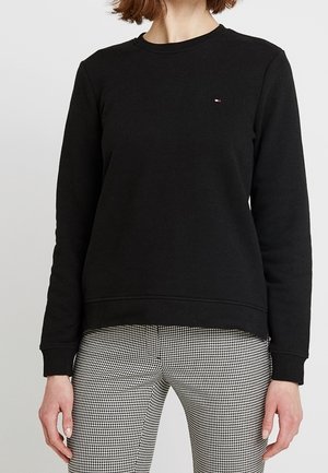 Femme portant un sweat-shirt noir à manches longues avec col rond et un pantalon à carreaux noir et blanc, debout, les bras détendus le long du corps.