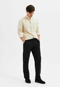 Light beige button-up shirt with long sleeves, paired with black trousers and black suede shoes. Minimalist design, no visible patterns.