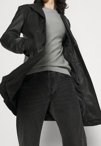 Manteau en cuir noir avec un col, doté d'une fermeture à boutons et d'une doublure intérieure lisse, superposé sur un pull gris et un jean noir.