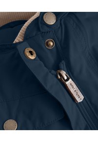 Marineblå jakke med en glatt, skinnende overflate. Har bronse hardware, trykknapper og en glidelås med «MINIATURE» merket på. Innvendig fôr er beige.